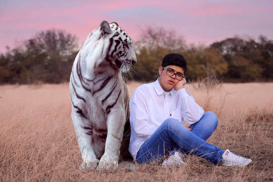 young man sitting in a field with a big tiger sitting next to him